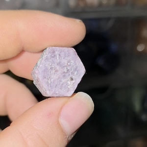 May include: A hexagonal, pale purple gemstone is held between two fingers. The gem has a rough, textured surface and a light colour. The gem is a natural mineral, possibly a ruby or sapphire.