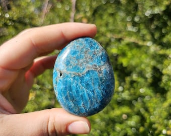 May include: A polished, oval-shaped blue apatite gemstone. The stone has a vibrant turquoise color with darker blue and gray veining. The surface is smooth and reflective, with a small natural cavity visible. The background is a blurred green.