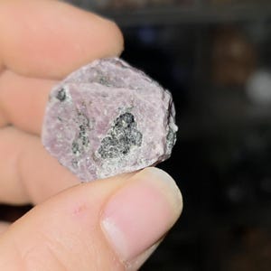 Könnte beinhalten: Ein roher, ungeschliffener Edelstein mit einem hellvioletten Farbton und dunkelgrauen Einschlüssen. Der Stein wird zwischen zwei Fingern gehalten, wodurch seine natürliche, unregelmäßige Form und Textur hervorgehoben werden. Es ist ein rohes Mineralexemplar.