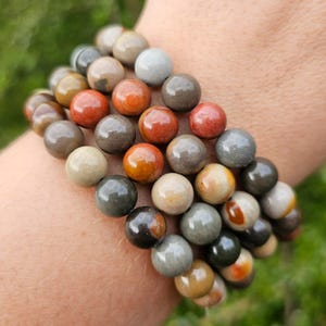 Könnte beinhalten: Ein Stapel Armbänder aus mehrfarbigen Jaspisperlen. Die Perlen sind rund und haben eine polierte Oberfläche. Die Armbänder sind auf dem Handgelenk einer Person gestapelt.