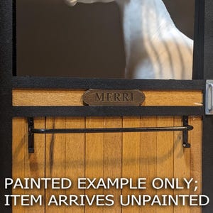 Puede incluir: Una puerta de establo en miniatura de madera con un marco negro y una barra de metal negra horizontal. Una pequeña placa ovalada con el texto MERRI está centrada en el panel superior. El texto PAINTED EXAMPLE ONLY; ITEM ARRIVES UNPAINTED es visible.