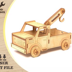 May include: A laser-cut wooden tow truck model. The truck features a detailed design with a lifting arm and hook. The wheels, cab, and bed are all visible. The text "WOOD TOOLBOX" and "LASER CUT FILE" are also visible.
