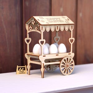 May include: A wooden Easter egg cart with a sign that reads "Happy Easter". The cart has a white background and is decorated with hearts and flowers. It is filled with six white eggs.