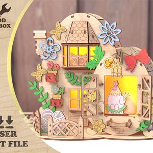 May include: A laser-cut wooden house with a whimsical design, featuring a hen illustration, flowers, butterflies, and a red bow. The house has a warm, inviting glow from the windows. The text "WOOD TOOLBOX" and "LASER CUT FILE" are visible.