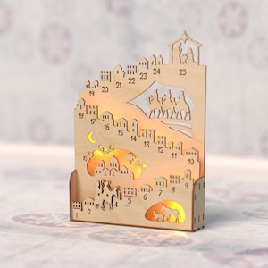 Op de afbeelding: Houten adventskalender met een stadsgezicht, uitgesneden ontwerpen en de nummers 1-25. De kalender heeft een halve maan, sterren en verlichte figuren. Het algehele ontwerp is lichtbruin.