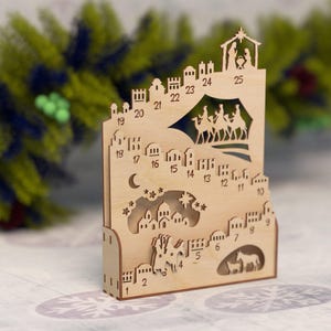 Op de afbeelding: Een houten adventskalender met een lasergesneden kerststal en genummerde ramen. De kalender toont een silhouet van een stad, een ster, een maan en de figuren van de drie wijzen. De nummers 1-25 zijn zichtbaar.