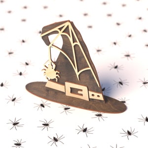 May include: A wooden witch's hat with a spider web design and a spider hanging from the web. The hat is sitting on a white surface with many black spiders scattered around it.
