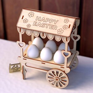 May include: A wooden Easter egg stand with a sign that reads "Happy Easter". The stand has a white painted finish and is decorated with hearts and flowers. It holds six white eggs.