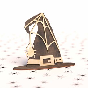 May include: A wooden witch's hat with a spider web design and a small spider hanging from the brim. The hat is brown with a black belt and is sitting on a white background with small black spiders scattered around it.
