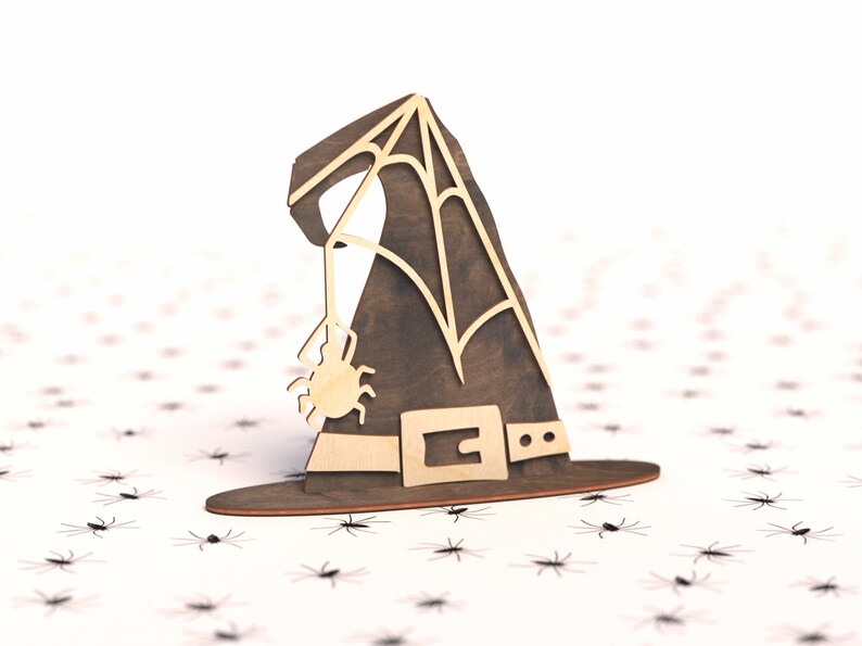 May include: A wooden witch's hat with a spider web design and a small spider hanging from the brim. The hat is brown with a black belt and is sitting on a white background with small black spiders scattered around it.