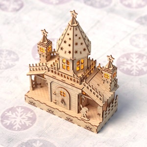 May include: A wooden Christmas house with a light brown finish and a star on top. The house has a porch, windows, and a door. The house is lit up with warm white lights.