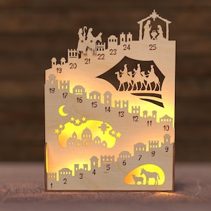 Op de afbeelding: Houten kerst adventskalender met lasergesneden silhouetten van stad en kerststal. Voorzien van de nummers 1-25, een maan, sterren en een gloeiend interieur. Een decoratief vakantieartikel.