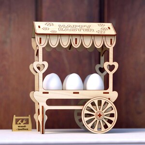 May include: Wooden Easter egg stand shaped like a cart with a sign that reads "Happy Easter". The cart has a white canopy and three white eggs inside.