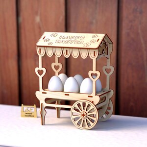 May include: A wooden Easter egg cart with a sign that reads "Happy Easter". The cart has four wheels and is decorated with hearts. It is holding five white eggs.