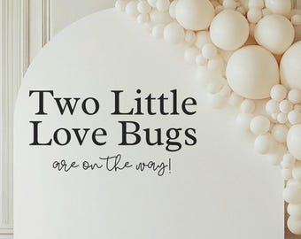 May include: A white arch backdrop with the text "Two Little Love Bugs are on the way!" is decorated with white balloons. A wooden rocking horse stands in front of the backdrop, creating a charming scene for a baby shower or gender reveal.