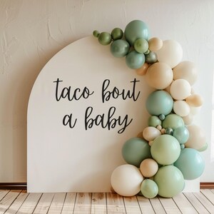 May include: A white arch with the text "taco bout a baby" written in black. The arch is decorated with a garland of green, beige, and white balloons.