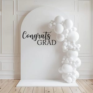 May include: White balloon arch with the text "Congrats Grad" on a white backdrop. The balloons are arranged in a cascading pattern, creating a festive and celebratory atmosphere.