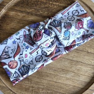 May include: White headband with a repeating pattern of wizarding-themed illustrations. The design includes a lightning bolt, a potion bottle, a wand, and a sorting hat. The headband is knotted at the top.