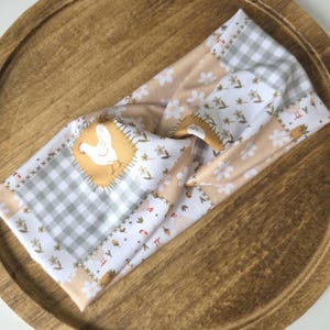 May include: A fabric headband with a patchwork design featuring a chicken, floral, and gingham patterns in white, tan, and gray. The headband is twisted at the center and rests on a wooden tray.