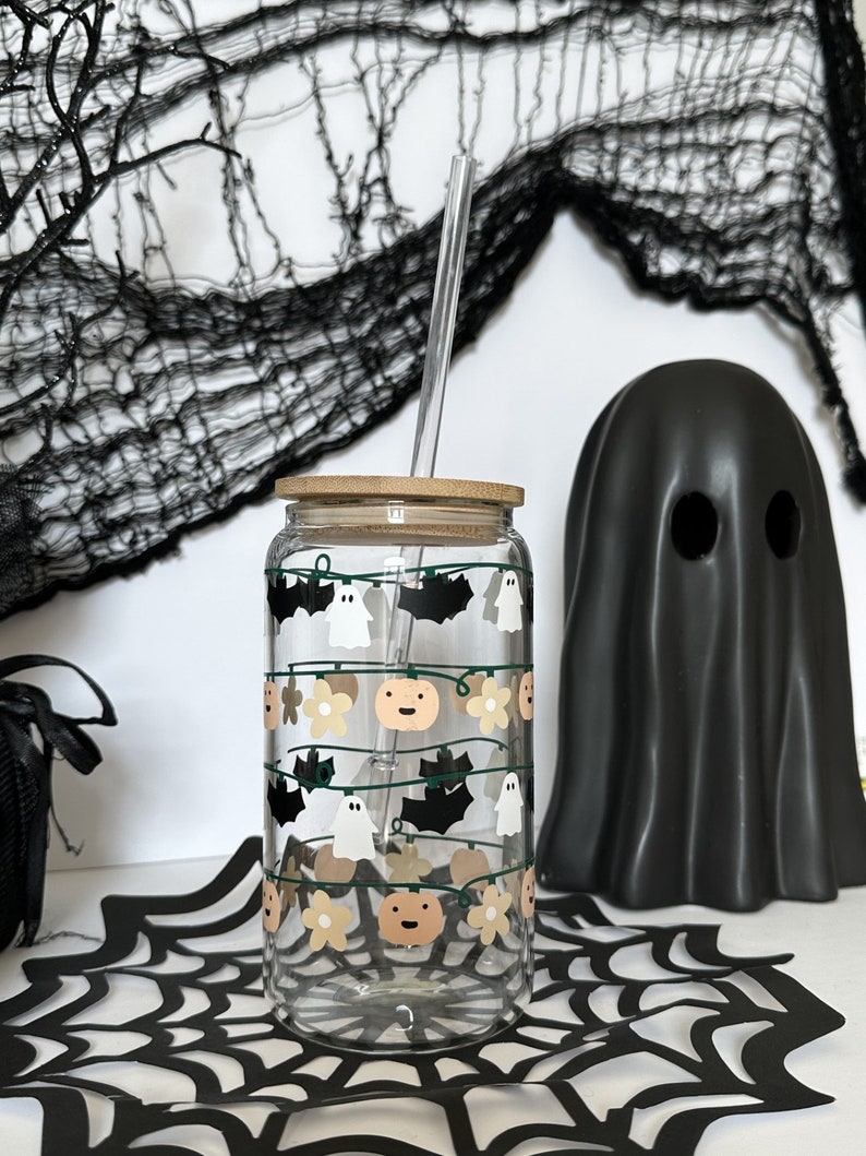 May include: A clear glass reusable water bottle with a bamboo lid and a straw. The bottle has a Halloween themed design with ghosts, bats, pumpkins, and flowers.