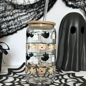 May include: A clear glass tumbler with a bamboo lid and a straw. The tumbler has a Halloween-themed design with ghosts, bats, pumpkins, and flowers. The tumbler is sitting on a black spiderweb-patterned surface.