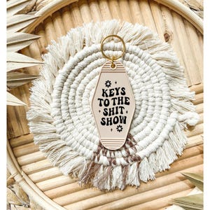 Peut inclure: Un porte-clés de motel beige avec un pompon noir et le texte "Keys to the Shit Show" en lettres noires.