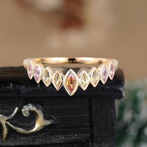 May include: A gold ring featuring a row of marquise-cut gemstones in shades of pink, yellow, and orange. The ring is set against a dark, textured surface, with a blurred background.