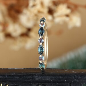 May include: A delicate gold band ring with a row of oval-shaped blue gemstones. The ring is set on a black surface.