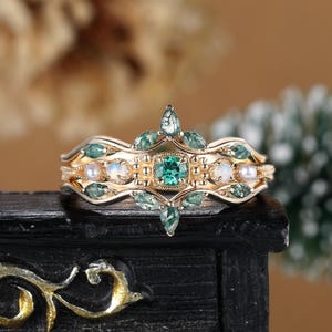 Op de afbeelding: Gouden ring met een groene edelsteen omringd door kleinere groene edelstenen en witte parels. De ring heeft een delicaat, ingewikkeld ontwerp.