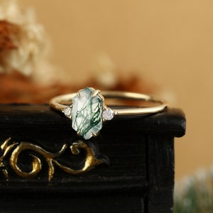 Pu&ograve; includere: Un anello d'oro con una pietra preziosa di agata verde muschio e due piccoli diamanti. L'anello &egrave; posto su una superficie nera.