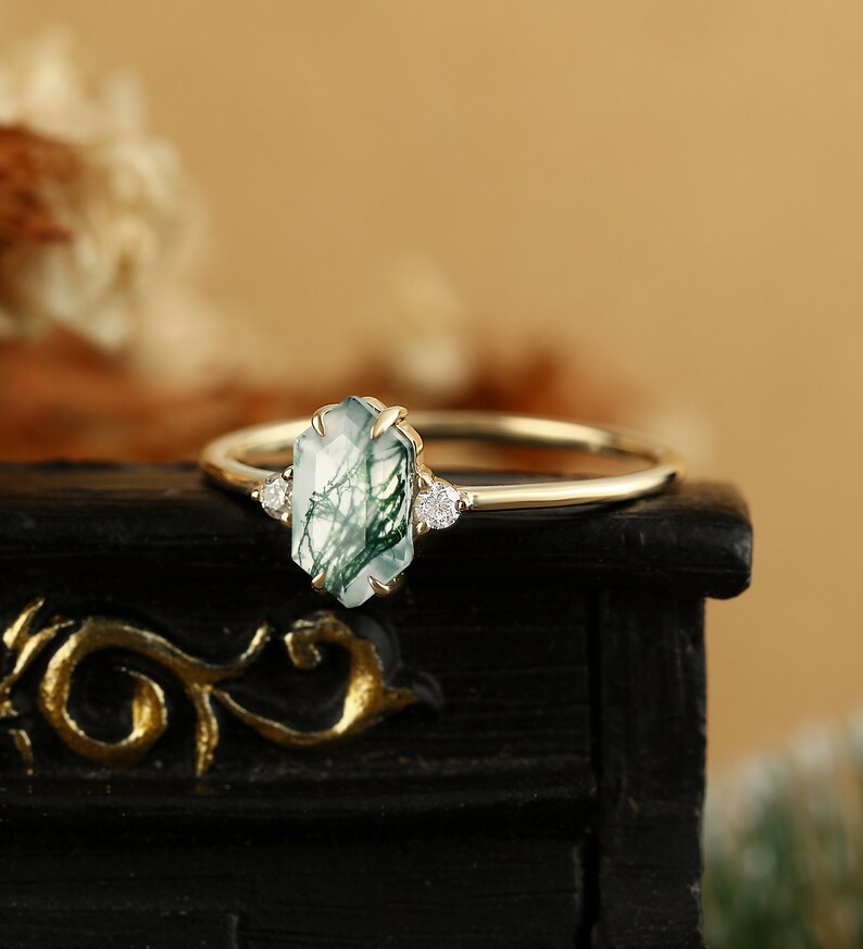 Pu&ograve; includere: Un anello d'oro con una pietra preziosa di agata verde muschio e due piccoli diamanti. L'anello &egrave; posto su una superficie nera.