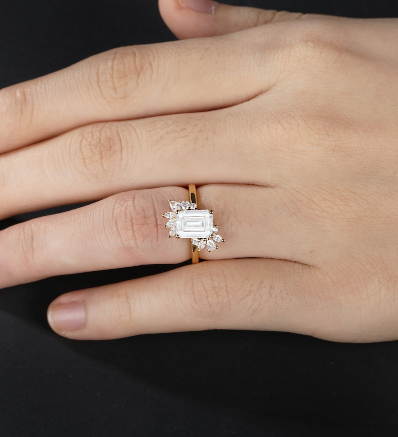 K&ouml;nnte beinhalten: Ein goldener Verlobungsring mit einem gro&szlig;en, rechteckigen Brillanten im Smaragdschliff und kleineren runden und birnenf&ouml;rmigen Akzent-Steinen.