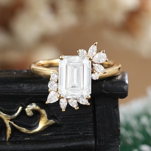 K&ouml;nnte beinhalten: Ein goldener Verlobungsring mit einem gro&szlig;en Smaragdschliff-Diamanten und kleineren Diamanten, die in einem floralen Design gesetzt sind.