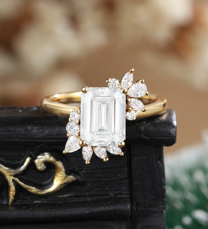 K&ouml;nnte beinhalten: Ein goldener Verlobungsring mit einem gro&szlig;en Smaragdschliff-Diamanten und kleineren Diamanten, die in einem floralen Design gesetzt sind.