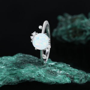May include: A silver ring featuring a white opal gemstone encircled by smaller clear gemstones. The ring is displayed on a green rock.