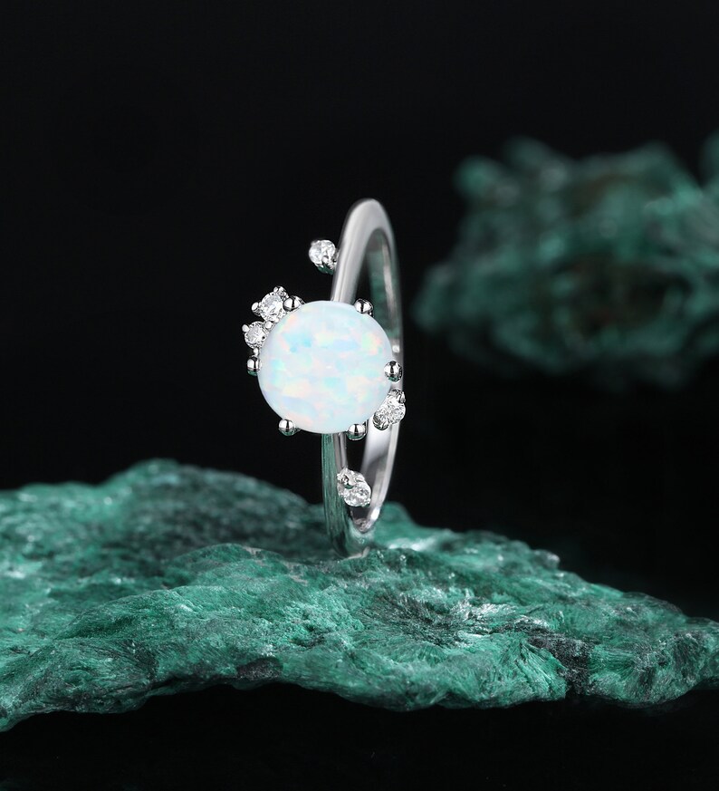 May include: A silver ring featuring a white opal gemstone encircled by smaller clear gemstones. The ring is displayed on a green rock.