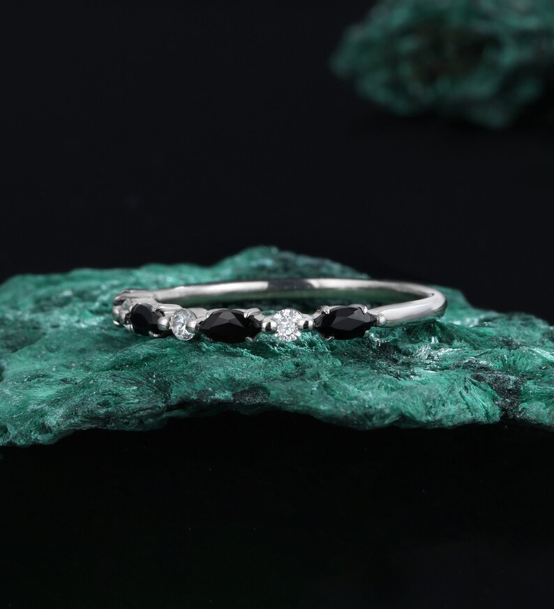 May include: A delicate silver band ring with a row of black and white gemstones. The ring is set on a green rock.