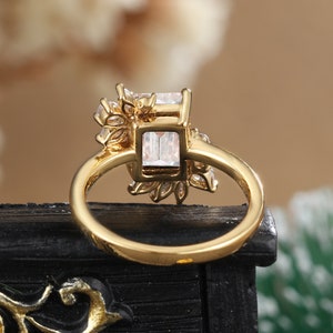 K&ouml;nnte beinhalten: Goldring mit einem gro&szlig;en quadratischen, klaren Stein, der von kleineren klaren Steinen umgeben ist. Der Ring liegt auf einer schwarzen Oberfl&auml;che.