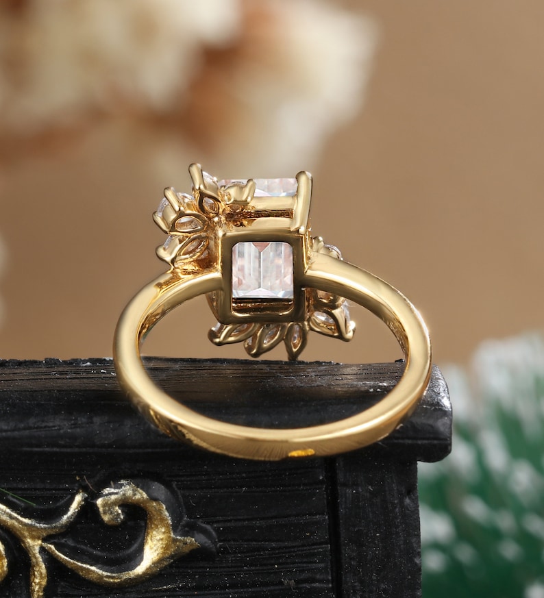 K&ouml;nnte beinhalten: Goldring mit einem gro&szlig;en quadratischen, klaren Stein, der von kleineren klaren Steinen umgeben ist. Der Ring liegt auf einer schwarzen Oberfl&auml;che.