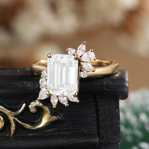 K&ouml;nnte beinhalten: Ein goldener Verlobungsring mit einem gro&szlig;en Smaragdschliff-Diamanten und kleineren Diamanten, die in einem floralen Design gesetzt sind.