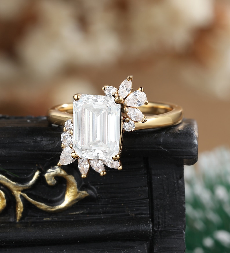 K&ouml;nnte beinhalten: Ein goldener Verlobungsring mit einem gro&szlig;en Smaragdschliff-Diamanten und kleineren Diamanten, die in einem floralen Design gesetzt sind.