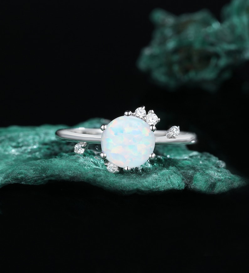 May include: A silver ring featuring a white opal gemstone and small clear cubic zirconia stones. The ring is displayed against a green backdrop.