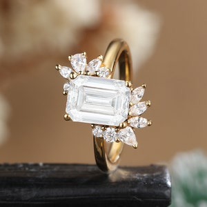 K&ouml;nnte beinhalten: Ein goldener Ring mit einem gro&szlig;en Smaragdschliff-Diamanten und kleineren Diamanten, die in einem floralen Design gesetzt sind.