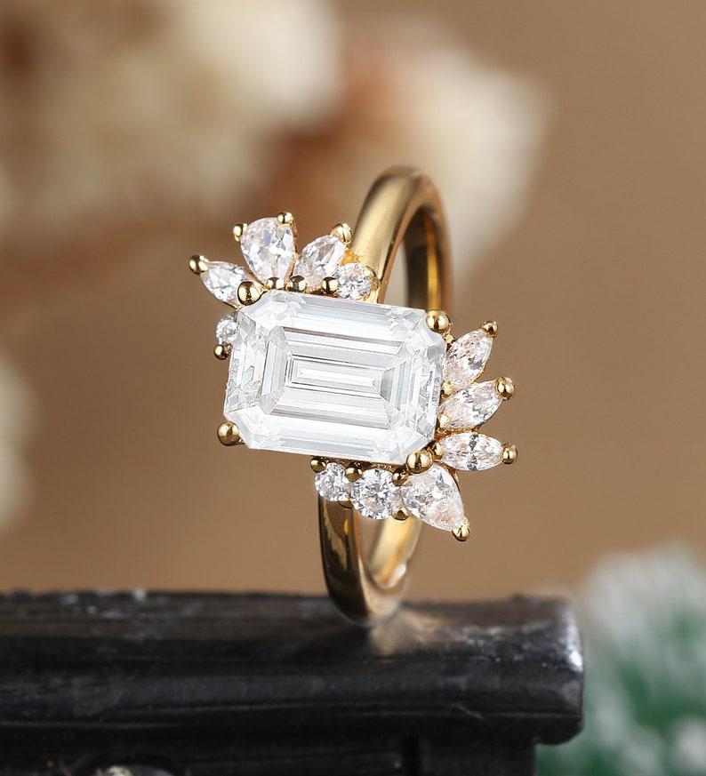 K&ouml;nnte beinhalten: Ein goldener Ring mit einem gro&szlig;en Smaragdschliff-Diamanten und kleineren Diamanten, die in einem floralen Design gesetzt sind.