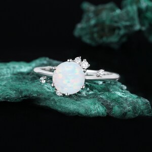 May include: A silver ring featuring a white opal gemstone and small clear cubic zirconia stones. The ring is displayed against a green background.