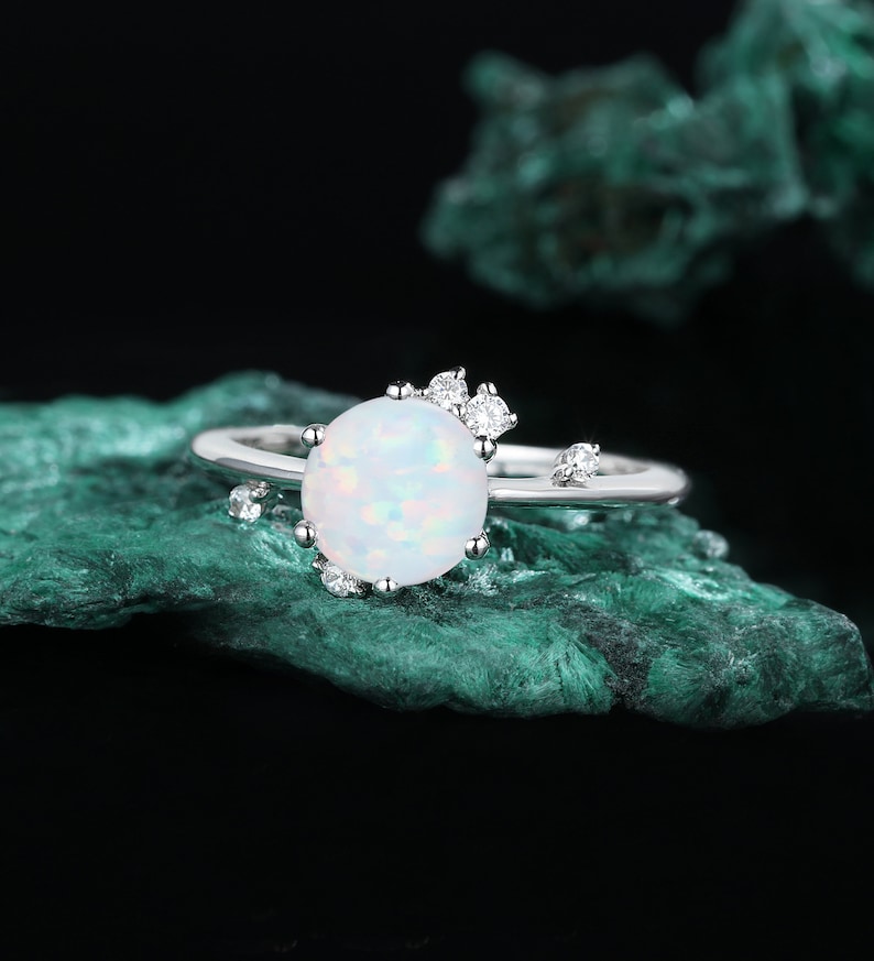 May include: A silver ring featuring a white opal gemstone and small clear cubic zirconia stones. The ring is displayed against a green background.
