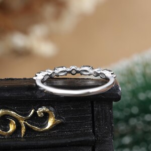 May include: A silver band ring with a row of small, clear, oval-shaped stones. The ring is set on a black surface.