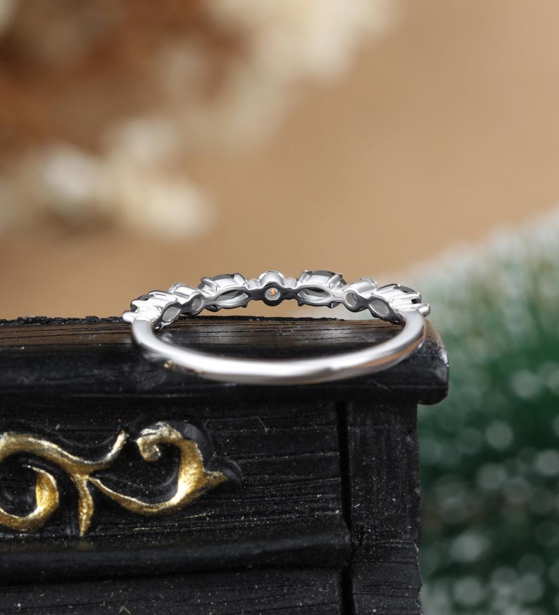 May include: A silver band ring with a row of small, clear, oval-shaped stones. The ring is set on a black surface.
