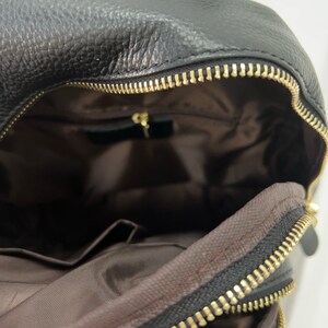 May include: A black leather backpack with a gold zipper. The backpack is open and the inside is lined with brown fabric. There is a small pocket on the inside of the backpack.