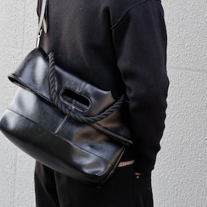 Puede incluir: Un bolso de hombro negro con acabado liso, diseño de solapa plegada y un asa gruesa de cuerda trenzada integrada en la estructura. El bolso incluye un asa recortada rectangular y una correa de hombro larga y ajustable para llevarlo.